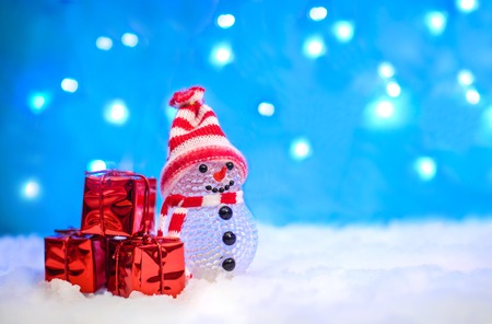 Christmas holiday picture with a cute little snowman in a red knitted hat and gifts in a red package in the snow on a blue background with lightsの写真素材