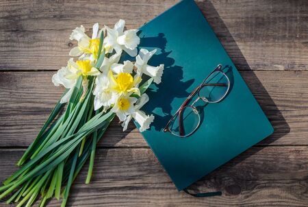 A bouquet of white and yellow daffodils, a closed book and glasses on a rustic wooden backgroundの写真素材