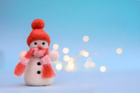 Christmas toy felted wool snowman in a red winter hat on a blue background with bokeh, copy space
