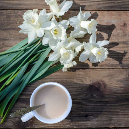 A beautiful bouquet of spring white flowers daffodils and a cup of coffee with milk on a wooden backgroundの写真素材