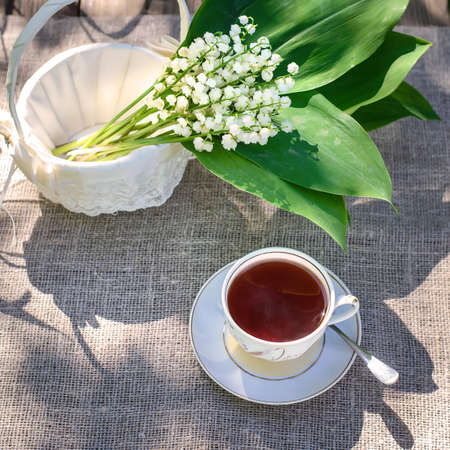Spring bouquet of lilies of the valley with green leaves in a beautiful white basket with lace and a cup of teaの写真素材