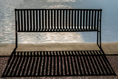 back of an empty metal bench by a reflective lake with sun on water and shadow behind benchの写真素材