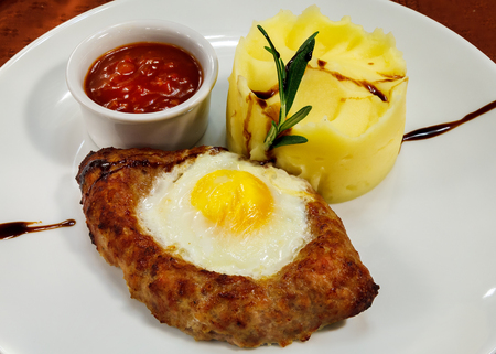 Baked patty in the shape of a boat with egg on top, mashed potatoes and spicy tomato sauce on a white plateの写真素材