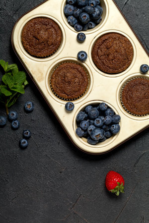 Freshly baked homemade cupcakes with fresh blueberries, strawberries and mint leaves on black background. Homemade baking concept. Soft focusの写真素材