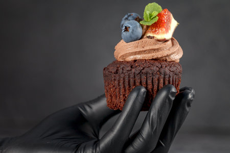 Muffin in hand. Chocolate cupcake. Protective glove. Muffin with cream and fruit on a dark backgroundの写真素材