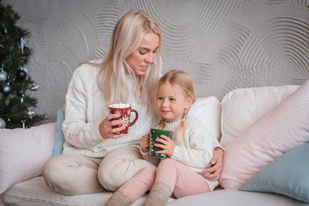 Young beautiful blonde woman hugs her little daughter on the couch with mugs of warm drink. Mother...
