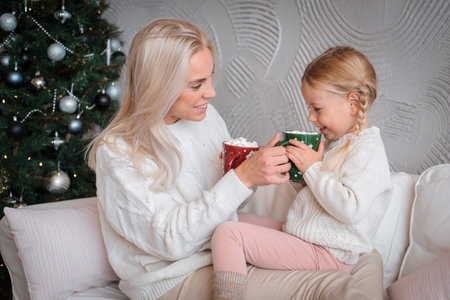 Young beautiful blonde woman hugs her little daughter on the couch with mugs of warm drink. Mother...