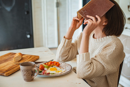 young girl is reading an old book at breakfast. Close-up hands and dinner table setting. Country style. Scrambled eggs and fresh vegetables and reading an interesting book in the morningの写真素材