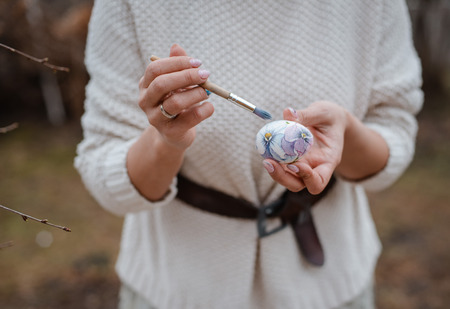 Close-up of a girl's hand painting an Easter egg in an outdoor garden. Farm. Rustic style. Willow spring bloom. Flowers cookies glass jar. Easterの写真素材
