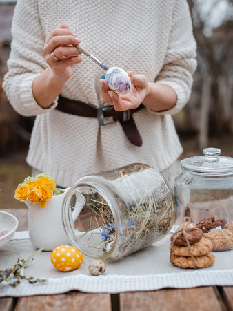 Close-up of the girl's hand paints an Easter egg in the garden at the laid holiday table outdoors. Farm. Rustic style. Willow spring bloom. Flowers cookies glass jar. Easterの写真素材