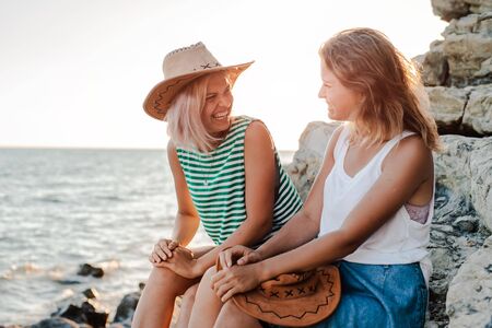 Two young cheerful women in hipsters hats on a rock on the coast of the sea. Summer landscape with girl, sea, Islands and orange sunlight. Travel and tourism with a friend. Twin sister, familyの写真素材