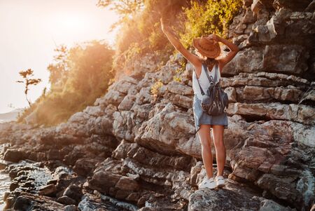 A young hipster woman hat and a rukzak with her hands up, standing on top of a cliff and looking sea at sunset. Summer landscape with girl, sea, Islands and orange sunlight. Travel and tourism.の写真素材