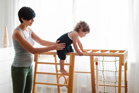 Mom supports a child aged 1.5 years engaged in a home childrens wooden sports complex with a slide. Early development of physical activity of the child.の写真素材