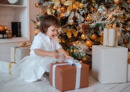 Pretty little girl opens a Christmas present. Portrait of happy kid on Christmas eve. Cozy living room with decorated fir treeの写真素材