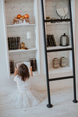 Girl child preschooler in a white fluffy dress sitting on the floor and reading an open book. The view from the top. White interior, shelves.の写真素材