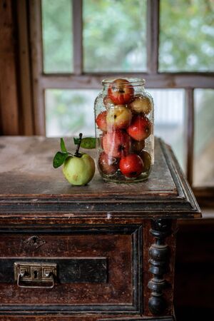 Fresh harvest of ripe and healthy farm apples in a glass jar, in a basket. Still life autumn rustic by the window and an old chest of drawers on a dark background.の写真素材