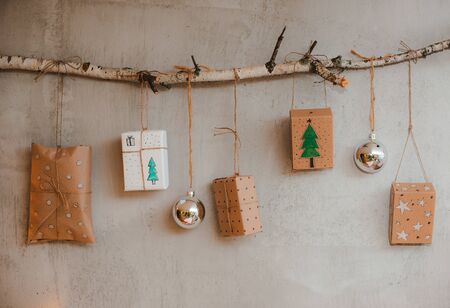 Christmas gifts Packed with Kraft paper and hand made decorations hang on a rope tied to a stick against a gray concrete wall. Concept of holiday and new year 2020 at homeの写真素材