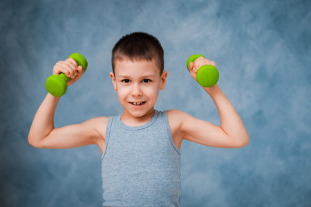 Cry sports baby boy emotionally shows his arm bicep muscle strength with dumbbells on a grey blue...