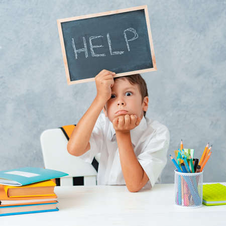 Sad tired frustrated schoolboy boy at the table and holding paper with word Help. Learning difficulties, education concept.の写真素材