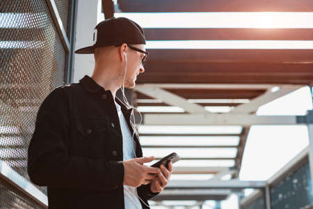 trendy stylish guy in a black cap and sunglasses with a smartphone and headphones on the background of an urban urban sunset. people, music, technology, leisure and lifestyleの写真素材