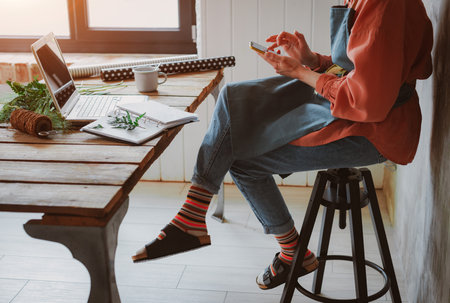 Fashionable stylish girl hipster florist in a work apron working at a laptop, looking at a smartphone check online stores for sales cyber monday technologyの写真素材