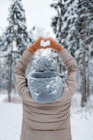 happy woman walking outside woods snowy winter day. woman mittens dressed fashionable gray stands with her back makes heart gesture with her hands against background snowy winter forestの写真素材
