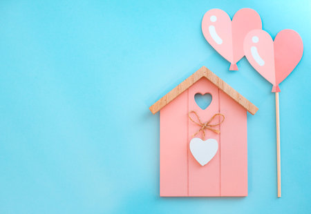 Miniature pink wooden flat toy house with hearts on blue background. Two heart-shaped balloons St. Valentines Day composition.の写真素材