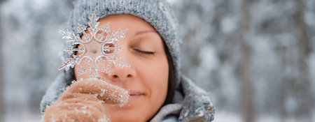 Portrait of young happy woman with eyes closed wearing knitted hat and mittens holding artificial snowflake in front of face, standing against winter snowy forest and falling snow, selective focusの写真素材