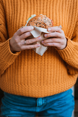 Healthy fast food. Cropped photo of unknown girl in orange sweater holding tasty looking hamburger...