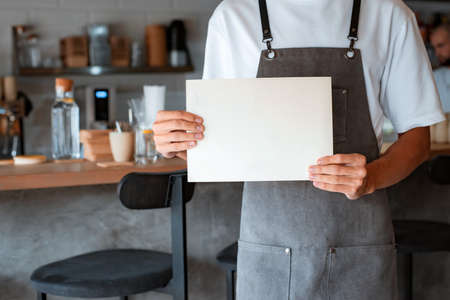 ropped photo of unknown man coffee shop owner or cafe worker holding blank sheet in hands while standing against background of wooden counter in coffeehouse, mockup for advertising. small businessの写真素材