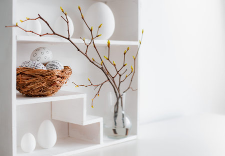 Home minimalist interior with Easter decor. Branches with budding buds in a glass vase on shelves in...
