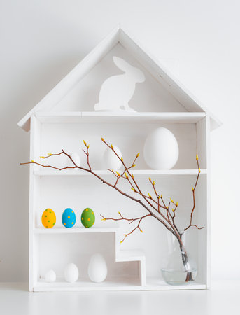 springtime Home minimalist interior with Easter decor. Branches with budding buds in a glass vase on shelves in the shape of a house, Easter eggs in a nest basket on a white background.の写真素材