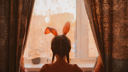 Children are expecting Easter. A girl child in bunny ears looks out the window at the sunset of the spring sun. The child dreams with hope for the future, open curtainの写真素材