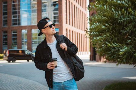 Portrait of a stylish hipster guy in sunglasses with a backpack in the city against the background of a modern city building. The concept of walking, traveling, leisureの写真素材