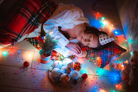 Cozy photo of lonely peacefully sleeping woman covered with blanket and led lights on parquet floor under Christmas tree at home next to festive dining table. Female resting after New Year partyの写真素材