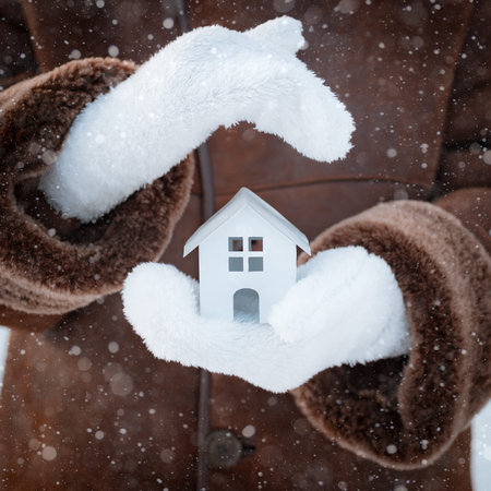 small model of a house in wool gloves on the street in winter against the background of snowの写真素材