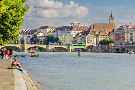 Basel city- A bridge spans Rhein river with a city in the background. The bridge is a stone...