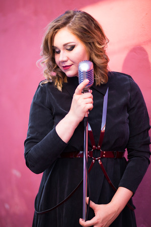 Beautiful sexy woman in dark velvet dress singing with the retro style microphone on pink backgroundの写真素材