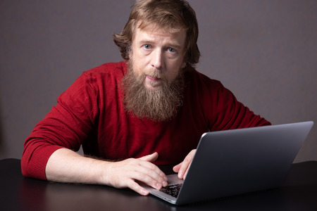 smiling bearded man wearing jumper red working with notebook in office looking into the cameraの写真素材