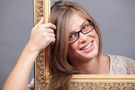 young woman in eyeglasses looking through wooden golden frame on grey backgroundの写真素材