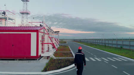 An oil and gas worker walks past fuel tanks to check the tightness and fuel level in a fuel warehouse. An employee in an orange helmet and black special clothing.の写真素材