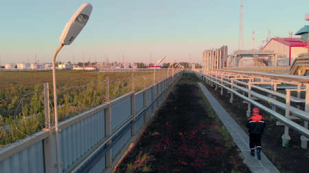 A drone flies behind an employee in an industrial area behind a fence with barbed wire, pipes with oil and gas pass from the employees right, in a protective casing.の写真素材