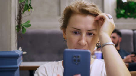 A blonde woman straightens her hair using a smartphone camera as a mirror. A woman is sitting in a cafe waiting for a meeting with a man.の写真素材
