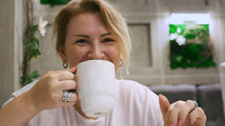 The girl drinks coffee from a white mug, thinks for a second and looks cheerfully at the frame and winks with a smile. Cheerful morning mood.の写真素材