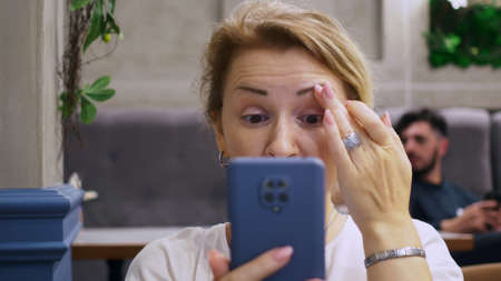 A blonde woman straightens her hair using a smartphone camera as a mirror. A woman is sitting in a cafe waiting for a meeting with a man.の写真素材