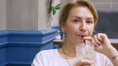 A blonde woman in a cafe is happy to drink a chocolate cocktail from a glass glass through a straw. The woman in the restaurant is brooding and dreamy.の写真素材