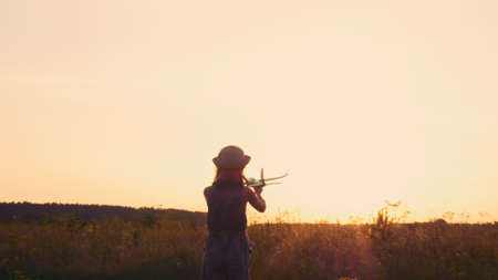 Silhouette of a teenage girl in a hat who plays with an airplane in a field in the rays of the setting sun. The girl is wearing a hat. Living in a dream and a village.の写真素材
