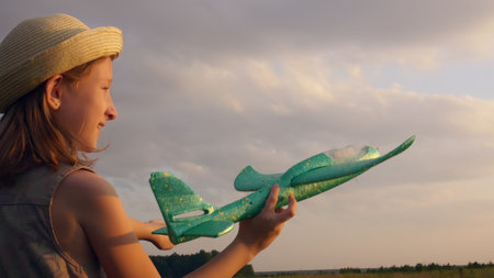 A beautiful blonde teenage girl in a straw hat in the rays of the setting sun plays with an airplane on a meadow. Happy childhood in dreams and an imaginary world.の写真素材