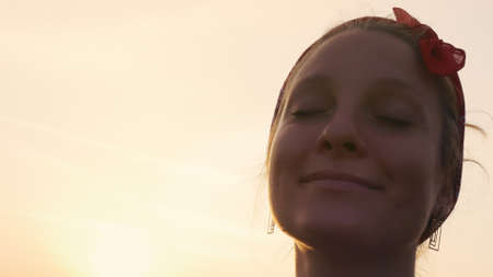 A beautiful girl in the rays of the setting sun is enjoying a moment in eternity. A blonde with a red scarf on her head, closing her eyes, dreams of happiness.の写真素材