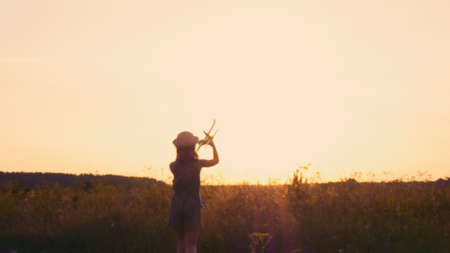 Silhouette of a teenage girl in a hat who plays with an airplane in a field in the rays of the setting sun. The girl is wearing a hat. Living in a dream and a village.の写真素材
